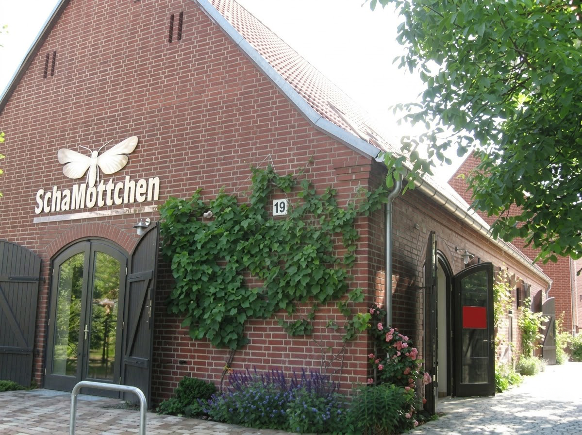 Backsteingebäude mit Schriftzug "SchaMöttchen", Schmetterlingslogo, offenen Türen und Kletterpflanzen.